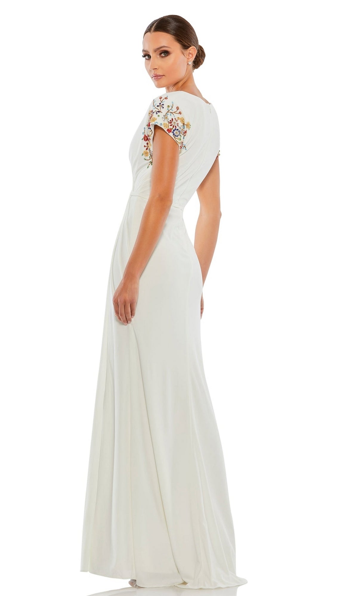 Woman in a white Mac Duggal 55709 Cap-Sleeve Long Formal Gown, featuring floral embroidery, stands and looks over her shoulder against a white background.